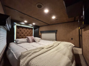 Cozy bedroom with a large bed, white bedding, and decorative pillows. The room&rsquo;s warm atmosphere feels like the inviting living quarters of a 5 horse trailer, featuring brown walls, a tufted headboard, ceiling lights, and window blinds. -Stillwater Trailer Sales