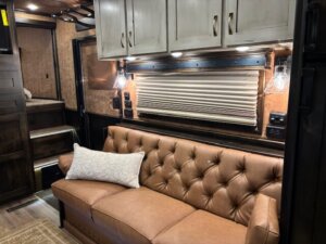 A brown leather tufted sofa with a white decorative pillow sits beneath a window with a closed pleated blind in this cozy Bloomer 5 Horse Trailer living quarters, featuring dark wood cabinets, built-in lighting, and carpeted steps in the background. -Stillwater Trailer Sales
