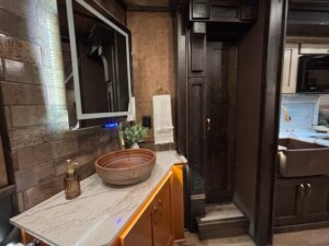 A modern bathroom with a vessel sink on a marble countertop, a wall-mounted mirror, brown cabinetry, and warm ambient lighting&mdash;perfect for relaxing in the Living Quarters of your Bloomer 5 Horse Side Load trailer. -Stillwater Trailer Sales