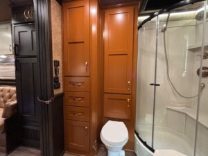 A compact bathroom with wooden cabinets, a white toilet, and a curved glass shower enclosure with a handheld showerhead. Dark brown finishes add warmth, making it ideal for side load horse trailer living quarters. -Stillwater Trailer Sales