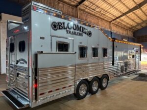 A large, shiny silver Bloomer side load horse trailer with multiple windows and three axles is parked inside a spacious warehouse with high ceilings and industrial lighting. -Stillwater Trailer Sales