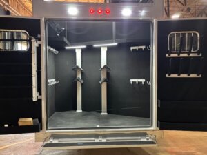 The image shows the open rear of a Bloomer horse trailer with a clean, empty tack room featuring multiple saddle racks, bridle hooks, and wall-mounted racks, all under bright interior lighting. -Stillwater Trailer Sales
