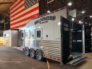 Large silver Bloomer horse trailer with living quarters, multiple windows, and open tack room doors, parked indoors under a large American flag. Sunlight shines through the building, highlighting the trailer&rsquo;s exterior. -Stillwater Trailer Sales