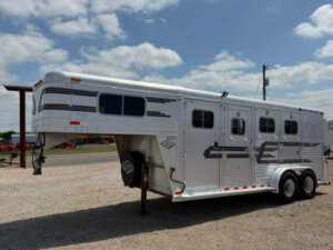 An Elite Horse Trailer, this silver gooseneck 3 horse trailer with black and red stripes features three windows and two axles. Parked on a gravel lot under a partly cloudy sky, it's positioned near a roadside. -Stillwater Trailer Sales