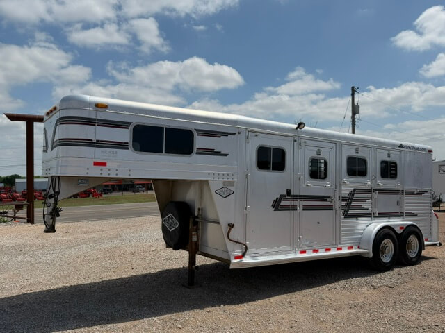 An Elite 3 Horse Trailer with a silver gooseneck, three side windows, and black and red striping is parked on a gravel lot under a partly cloudy sky. The trailer features two axles for added stability. -Stillwater Trailer Sales