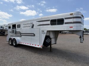 A silver Elite 3 Horse Trailer with red and black stripes is parked on a gravel lot under a partly cloudy sky. The side bears the quote, When all else fails, read the directions. -Stillwater Trailer Sales