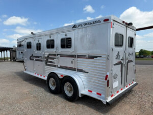 A silver Elite 3 Horse Trailer with three windows and dual axles is parked on gravel under a partly cloudy sky. The trailer features black and red stripes along with Elite Trailers branding near the top. -Stillwater Trailer Sales