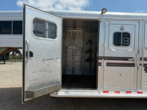 An open silver Elite 3 Horse Trailer with a door swung wide reveals an empty, metallic interior and saddle racks inside. Sunlight shines on the trailer, while another trailer is visible in the background. -Stillwater Trailer Sales