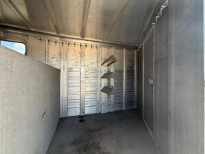 Interior of an empty Elite 3 Horse Trailer with three angled saddle racks mounted on one wall, a window to the left, a solid partition, and a door at the far end. The metallic floor and walls show some wear and stains. -Stillwater Trailer Sales