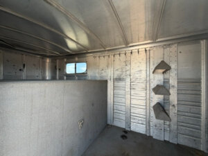 Interior of an empty Elite Horse Trailer 3 Horse Trailer with padded divider, small window on the left, and three angled saddle racks on the right wall. The metallic walls and ceiling show signs of use. -Stillwater Trailer Sales