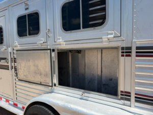 A close-up view of an Elite 3 Horse Trailer shows one lower compartment door open, revealing an empty storage area. The silver trailer features black-tinted windows and bold red and black horizontal stripes. -Stillwater Trailer Sales
