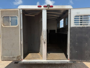 Wide open double doors reveal the empty interior of an Elite 3 Horse Trailer, with separate stalls, metal walls, and barred windows. Sunlight streams in, highlighting the clean, divided space inside. -Stillwater Trailer Sales