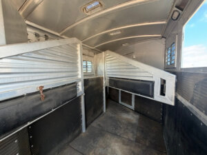 Interior of an empty Elite 3 Horse Trailer with metal dividers, padded side panels, and rubber flooring, designed to transport horses safely. Sunlight streams through vents and windows on the side and ceiling. -Stillwater Trailer Sales