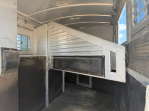 Interior of an Elite 3 Horse Trailer featuring padded dividers, metal walls, rubber flooring, and barred windows for ventilation, designed to safely transport animals. Bright light enters through the right-side windows. -Stillwater Trailer Sales