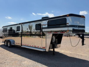 A large, shiny silver Hawk 2+1 GN horse trailer with black trim is parked on a gravel lot under a blue sky with a few clouds. The trailer features multiple windows and sits on double axles. -Stillwater Trailer Sales