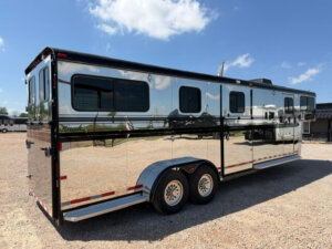 A shiny, silver Hawk 2+1 horse trailer with black trim and two axles is parked on gravel under a blue sky with scattered clouds. The trailer has several tinted windows and reflects its surroundings. -Stillwater Trailer Sales