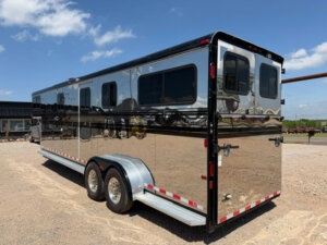 A shiny, silver and black Hawk 2+1 Horse Trailer with multiple windows is parked on a gravel lot under a blue sky with scattered clouds. -Stillwater Trailer Sales