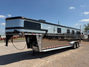 A large, shiny silver and black GN Horse Trailer with multiple windows is parked on gravel under a blue sky with scattered clouds. -Stillwater Trailer Sales