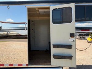 The image shows an open door of a Hawk 2+1 GN horse trailer, revealing the interior with saddle racks on the left wall. The trailer is parked on a gravel lot, with buildings, vehicles, and a clear sky in the background. -Stillwater Trailer Sales