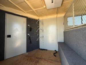 Interior of a Hawk 2+1 horse trailer utility room with a padded bench, wall hooks holding hangers and ropes, closed white door, gray carpeted walls, and a metal mesh divider on the right. -Stillwater Trailer Sales