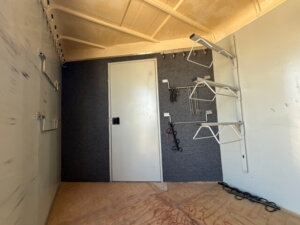 Interior of a storage room inside a GN horse trailer with a white door at the center, hooks and racks on the right wall, and a few tools and cables hanging. The floor is unfinished and the walls are painted light gray. -Stillwater Trailer Sales