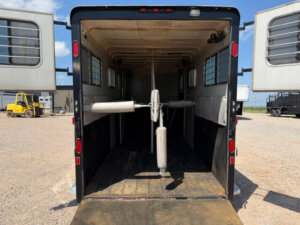 Open GN Horse Trailer with doors wide, padded dividers inside, and bright sunlight outside. The wooden-floored Hawk 2+1 is empty, with a yellow forklift and other vehicles visible in the background. -Stillwater Trailer Sales
