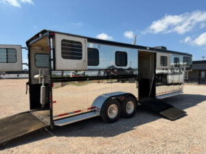 A shiny black and silver Hawk 2+1 horse trailer with barred windows, double axles, and two open ramps is parked on gravel beneath a blue sky with scattered clouds. -Stillwater Trailer Sales