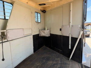 The interior of a GN horse trailer with padded walls, two small windows with bars, storage pouches, and a non-slip rubber floor; a door leads to another compartment. Sunlight shines in from outside. -Stillwater Trailer Sales