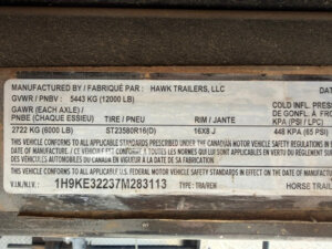 A metal label shows manufacturing details for a Hawk 2+1 GN horse trailer, including weight ratings, tire and rim size, and a VIN number. The text is in both English and French. -Stillwater Trailer Sales
