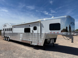 A large, silver and white Bloomer 5 Horse gooseneck trailer with multiple windows is parked on gravel under a clear blue sky. The trailer, labeled The Evolution on the front, features a spacious side load horse trailer design. -Stillwater Trailer Sales