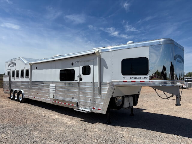 A large silver and white Bloomer gooseneck horse trailer with multiple windows, three axles, and side load capability is parked on gravel under a blue sky. The Evolution is written on the front side. 5 Horse capacity. -Stillwater Trailer Sales