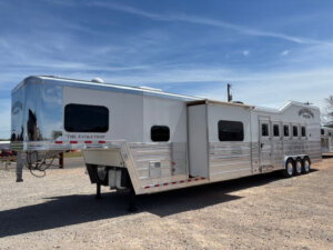 A large silver and white Bloomer 5 Horse gooseneck trailer with multiple windows and three axles is parked on a gravel lot under a blue sky. The Evolution is written on its side, highlighting its Outlaw conversion and convenient side load design. -Stillwater Trailer Sales