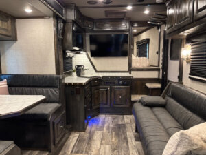 Interior of a modern Bloomer 5 Horse RV with dark wood cabinets, gray sofas, a side load design, built-in dining area with a marble-like table, kitchenette, flat-screen TV, and wood-style flooring. The space feels clean, cozy, and well-lit. -Stillwater Trailer Sales