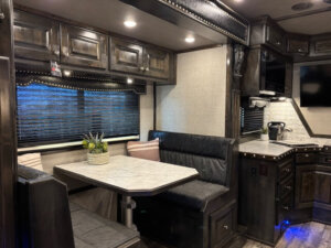 A modern RV interior inside a Bloomer 5 Horse Side Load, featuring a marble-look dining table with black cushioned bench seating, decorative pillows, a potted plant centerpiece, dark wood cabinets, and a compact Outlaw kitchen with TV and appliances. -Stillwater Trailer Sales