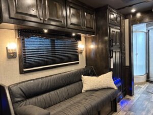 Modern Bloomer RV interior with dark wood cabinets, a gray leather sofa with a decorative pillow, wall sconces, window blinds, and a view of the bathroom with glass shower enclosure&mdash;perfect for your 5 Horse side load adventures. -Stillwater Trailer Sales