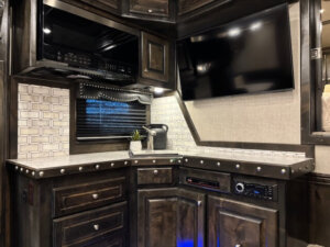 A modern RV kitchen corner inspired by 18' Outlaw design, featuring dark wood cabinets, a marble-look countertop, a small sink, coffee maker, potted plant, wall-mounted TV, and stainless appliances under bright lighting. -Stillwater Trailer Sales