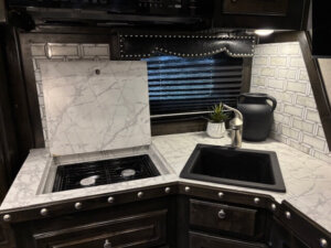 A modern Bloomer RV kitchen with marble countertops, a covered stovetop, a black sink, decorative backsplash tiles, a plant, and a black pitcher under a window with horizontal blinds. Perfect for relaxing after a day with your 5 Horse side load. -Stillwater Trailer Sales