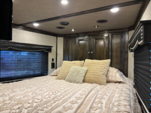 A cozy bedroom in a Bloomer 5 Horse RV features a neatly made bed with textured cream pillows and blanket, dark wooden cabinets above the headboard, and windows with black blinds on both sides. Warm overhead lights illuminate this Outlaw-inspired space. -Stillwater Trailer Sales