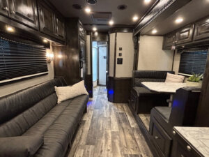 Interior of a modern 5 horse trailer RV with dark wood cabinets, a long black sofa with a pillow, a dining area featuring a marble-look table and black cushioned seats, and wood-look flooring. Doorway leads to a bathroom. -Stillwater Trailer Sales