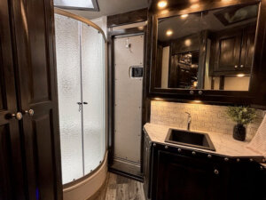 Modern RV bathroom in a Bloomer 5 Horse Trailer with a curved glass shower, black vanity with rectangular sink, marble-patterned countertop, mirrored cabinet, tiled backsplash, and a small plant decorating the counter. Side load design for convenience. -Stillwater Trailer Sales