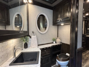 Modern bathroom with dark wood cabinets and a marble countertop features a square sink, oval mirror, decorative plants, a towel, and toilet in the corner. Neutral walls and geometric tile accents create a bloomer of style in the space. -Stillwater Trailer Sales