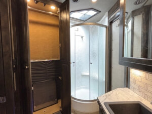 Modern RV bathroom in this Outlaw features a glass-enclosed rounded shower, open closet with shelves, marble countertop sink, mirrored cabinet, and geometric backsplash tiles. Ceiling lights and a skylight provide bright illumination. -Stillwater Trailer Sales