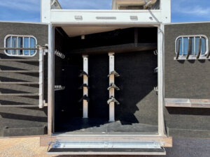 Open Bloomer horse trailer with rear doors wide, revealing an empty, carpeted tack room with multiple saddle racks and bridle hooks on the interior walls. Sunlight casts shadows across the space, highlighting its Outlaw interior. -Stillwater Trailer Sales