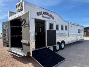 A large silver Bloomer 5 Horse trailer with multiple windows and open rear doors is parked on gravel. A black ramp extends to the ground, and tack storage is visible inside this stylish Outlaw edition. -Stillwater Trailer Sales