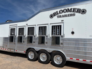 A large white Bloomer horse trailer with multiple windows and bars, this 5 horse side load Outlaw design is parked on a gravel surface under a blue sky. -Stillwater Trailer Sales