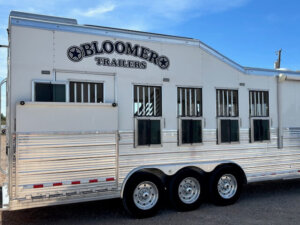 A white Bloomer horse trailer with an Outlaw conversion, five windows, metal bars, and three axles is pictured parked on gravel under a clear blue sky. -Stillwater Trailer Sales