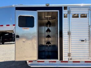 Open compartment of a silver Frontier horse trailer showing a clean, empty tack room with multiple black saddle racks inside. The door is open, and the 20' Stock Combo trailer is parked on gravel under a clear blue sky. -Stillwater Trailer Sales