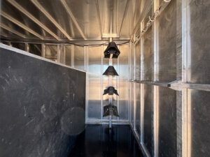 An interior view of a metallic, unfinished Frontier trailer or container with exposed studs and reflective walls, featuring three black saddle racks mounted vertically on a central pole&mdash;ideal for a 20' Stock Combo setup. -Stillwater Trailer Sales
