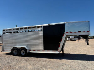 A silver Frontier stock combo gooseneck livestock trailer, 20' long with a black door, is parked on gravel under a clear blue sky. It features two axles with four wheels and red-and-white reflective tape along the edges. -Stillwater Trailer Sales