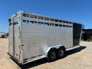 A silver Frontier stock trailer with slatted sides, two axles, and a black front door is parked on a gravel lot under a clear blue sky. This 20' Stock Combo offers durability and style for all your livestock needs. -Stillwater Trailer Sales